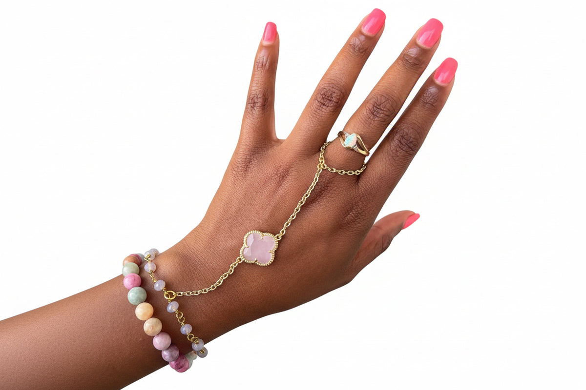 Hand wearing a gold bracelet with a pink clover charm and a beaded bracelet on a blurred background