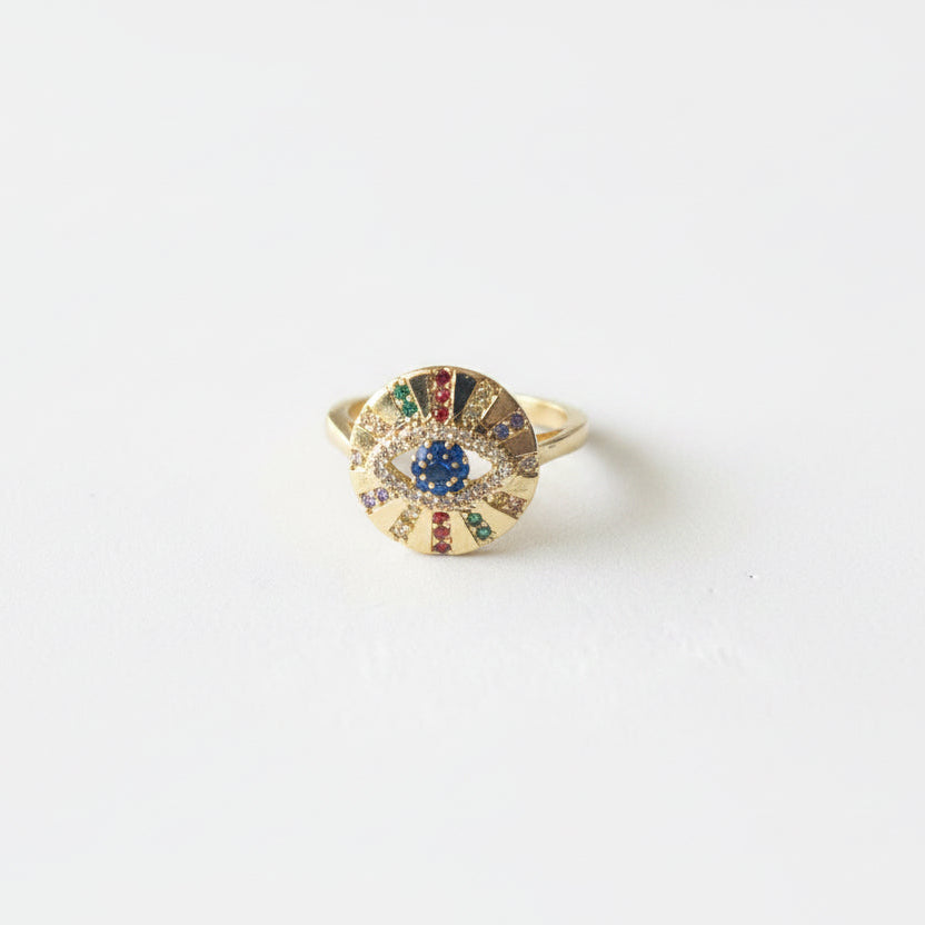 A gold ring with a circular sunburst design, featuring a central blue gemstone and smaller red, green, and yellow accent stones, set against a plain white background.