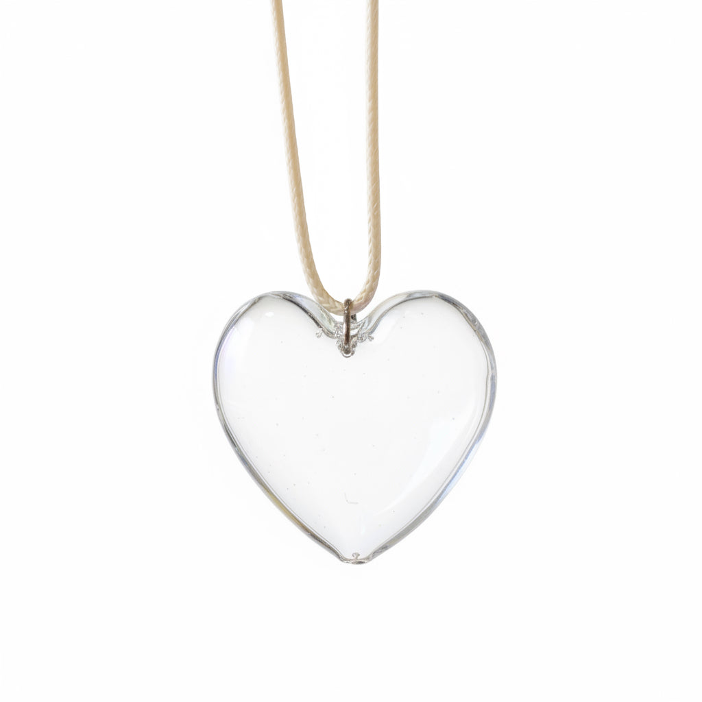 A heart-shaped glass pendant necklace with an iridescent, holographic finish, hanging from a thin beige cord. The background is a blurred outdoor patio setting with blue chairs, green artificial grass, and a glass door.